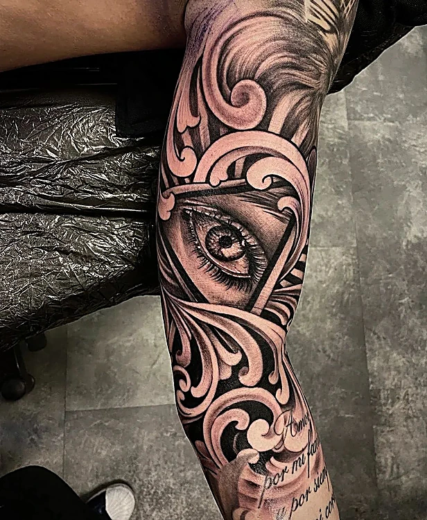 Ojo dentro de un triángulo rodeado de filigrana barroca en negro y gris, en el brazo