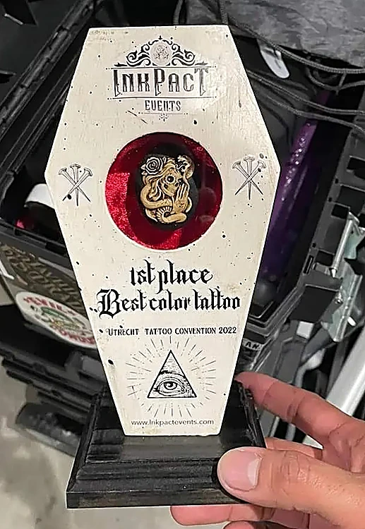 Trofeo blanco en forma de ataúd con una ventana de terciopelo rojo y el texto 1st place, Best color tattoo, Utrecht Tattoo Convention 2022, sostenido con la mano